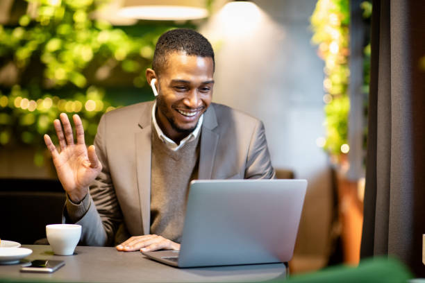 Video Call With Children. Happy afro businessman talking with family via laptop computer in cafe, waving hand, copyspace