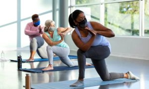 A multi-ethnic group of three women taking a yoga class, in a crescent lunge pose on knee with prayer hands. They are indoors, side by side, with the focus on the Caucasian woman in the middle. She is in her 50s. They are wearing protective face masks, trying to prevent the spread of coronavirus during the COVID-19 pandemic.